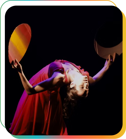 Bailarina em cena de espetáculo de dança contemporânea, vestindo figurino vermelho, com o corpo arqueado para trás e os braços abertos segurando adereços circulares coloridos, iluminada por luz cênica sobre fundo escuro.
